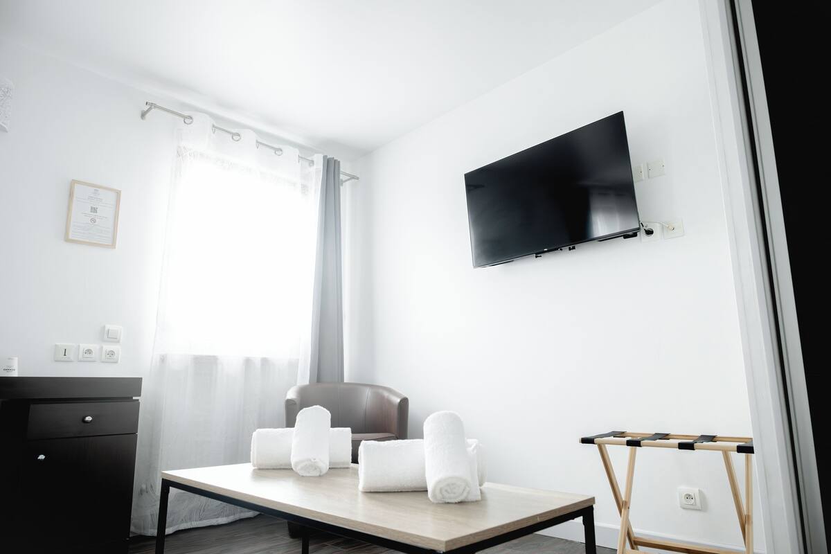 A modern hotel room with a flat-screen TV, a wooden table with towels, a chair, and a small desk with a mirror.