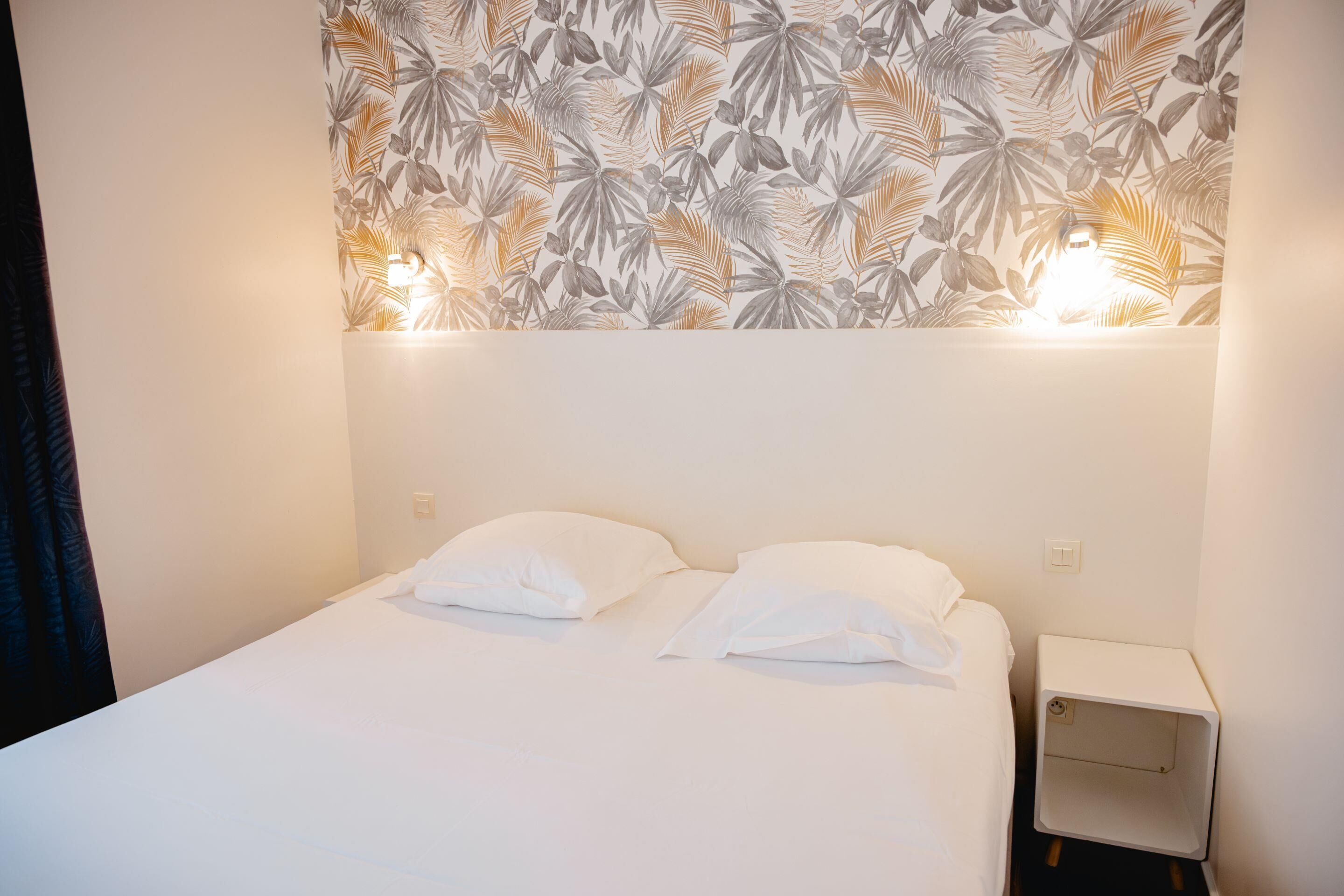 Una cama bien hecha con ropa blanca y almohadas, una mesita de noche pequeña y una pared con estampado de hojas.