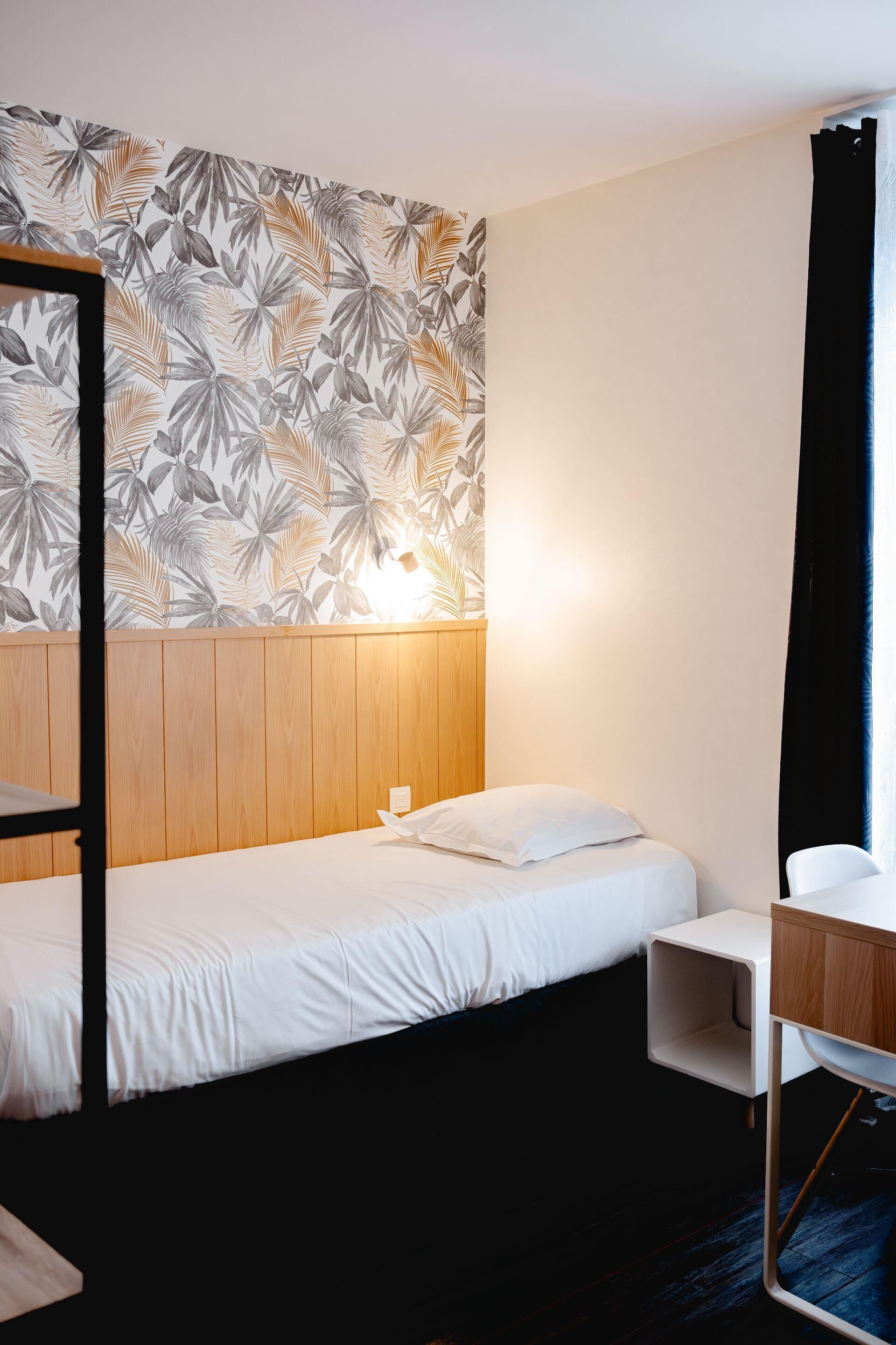 Een kamer met een stapelbed, een nachtkastje, een klein bureau en een muur met een tropisch bladmotief.