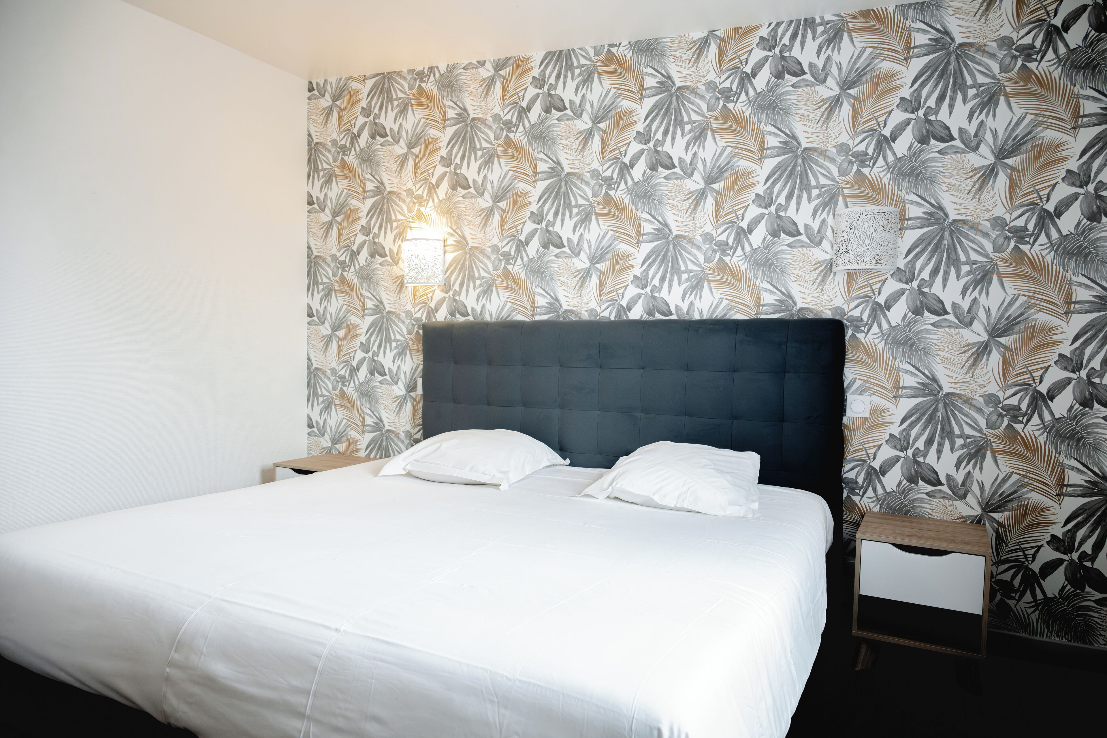 Une chambre à coucher avec un grand lit, une tête de lit sombre et un mur orné d’un motif de feuilles tropicales.