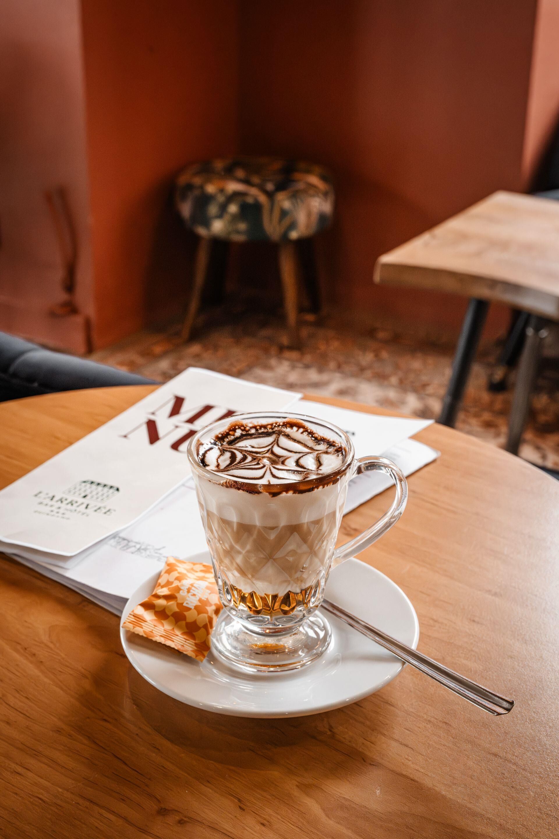 A clear glass coffee cup with latte art on a white saucer, placed on a wooden table with a magazine and a folded napkin beside it.