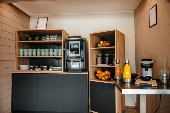 Una kitchenette con una cafetera, repisas con tazas y una tabla de cortar con naranjas.