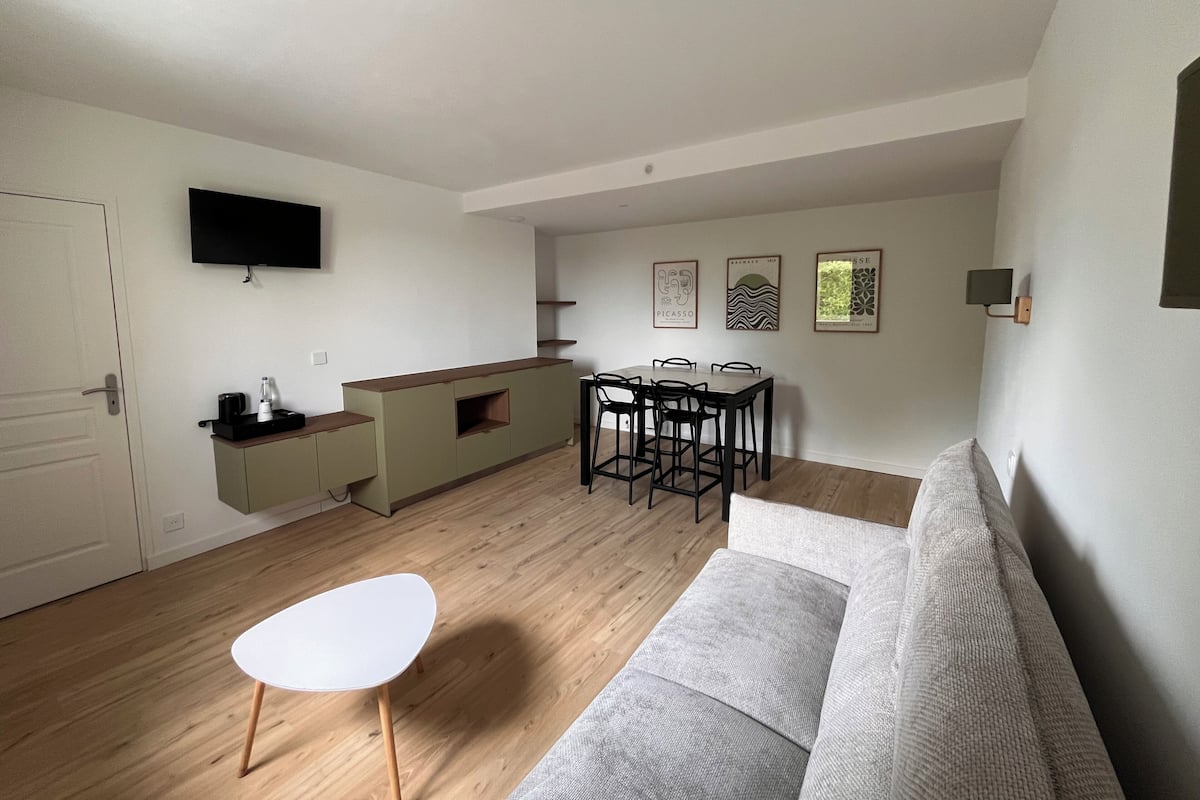 A modern living room with a sofa, a dining table with chairs, a kitchen area with a counter, and a flat-screen TV mounted on the wall.