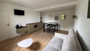 A modern living room with a sofa, a dining table with chairs, a kitchen area with a counter, and a flat-screen TV mounted on the wall.