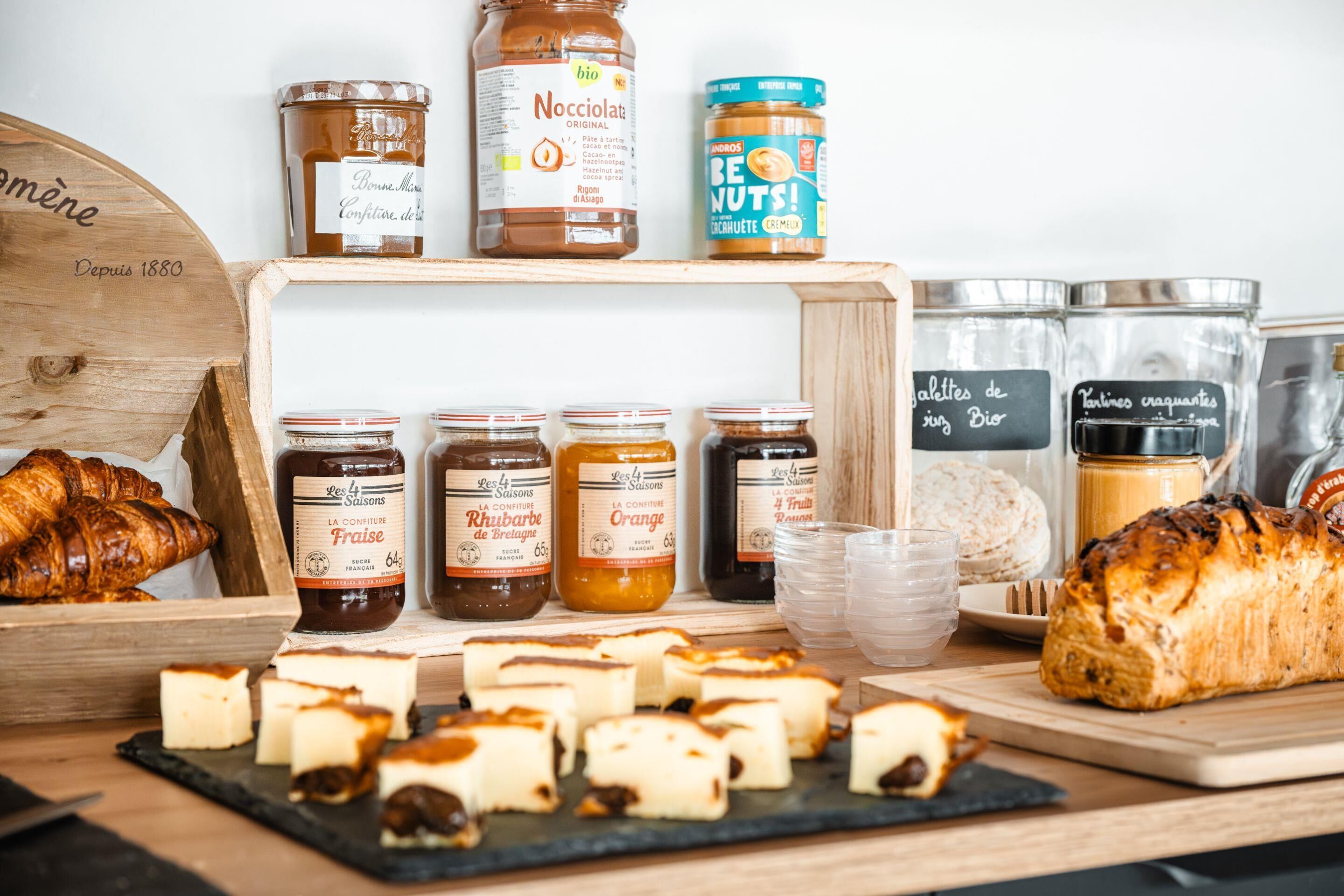 A variety of food items including jars of preserves, a loaf of bread, and a croissant on a wooden table.
