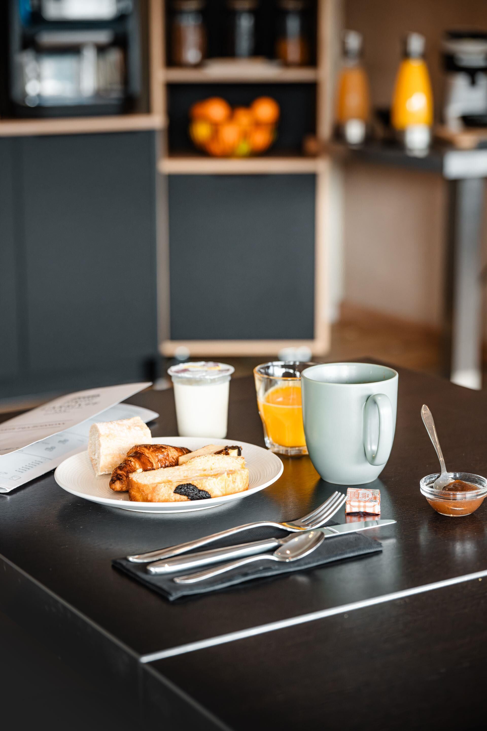 Une table mise pour le petit-déjeuner, avec une assiette de nourriture, une tasse, un verre de jus d’orange et un petit bol de confiture.
