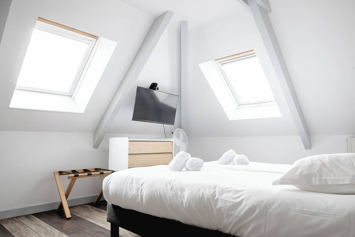 A modern bedroom with a sloped ceiling, two skylights, a bed with white bedding, a bedside table, a TV, and a fan.