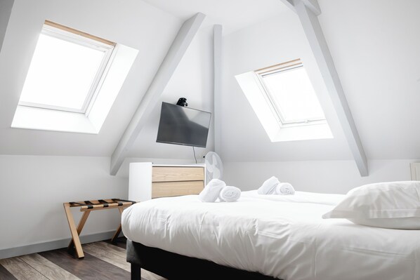 Un dormitorio moderno con techo inclinado, dos tragaluces, una cama con ropa de cama blanca, una mesita de noche, un televisor y un ventilador.