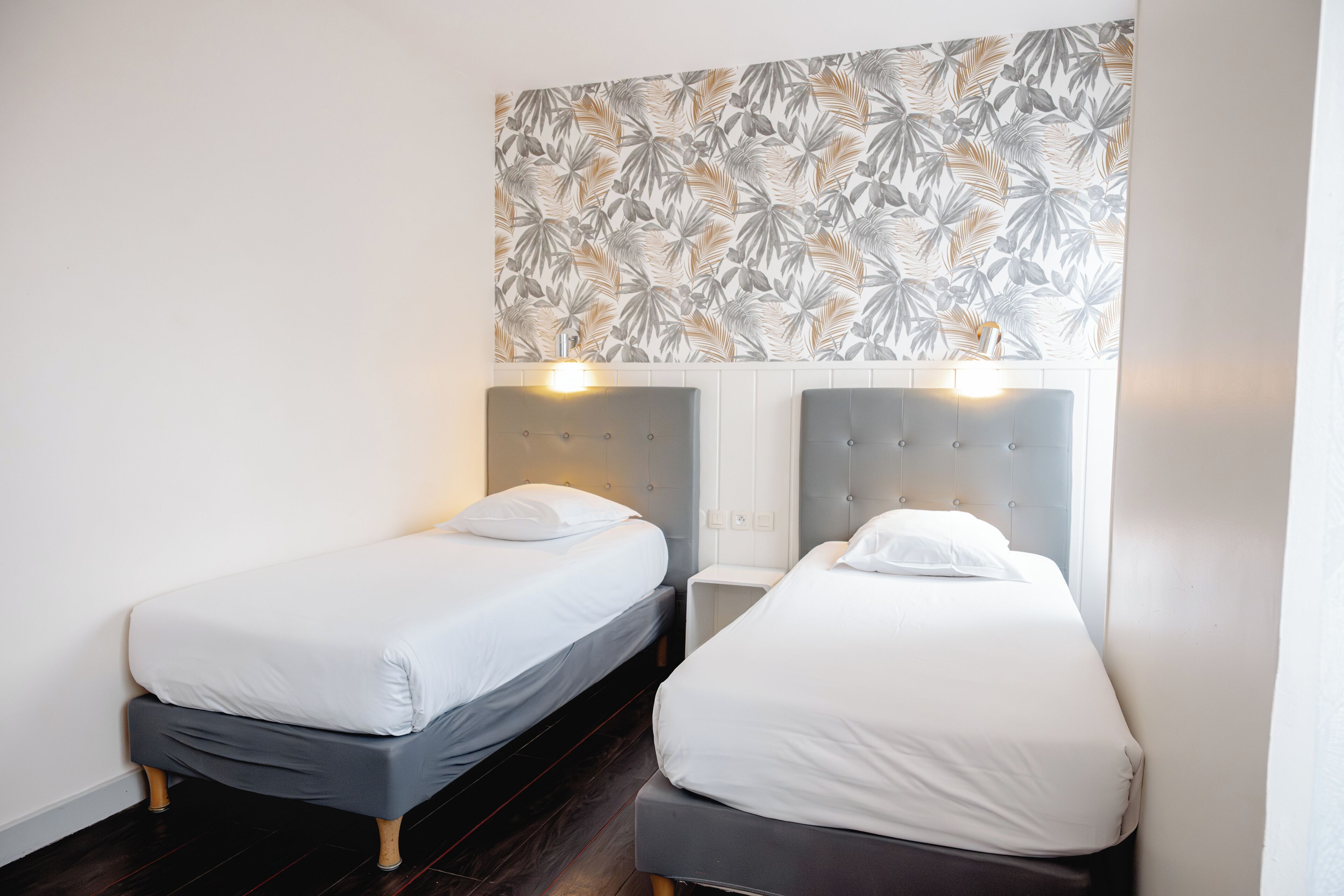 Two single beds with grey headboards and white bedding in a room with a tropical leaf-patterned wall.