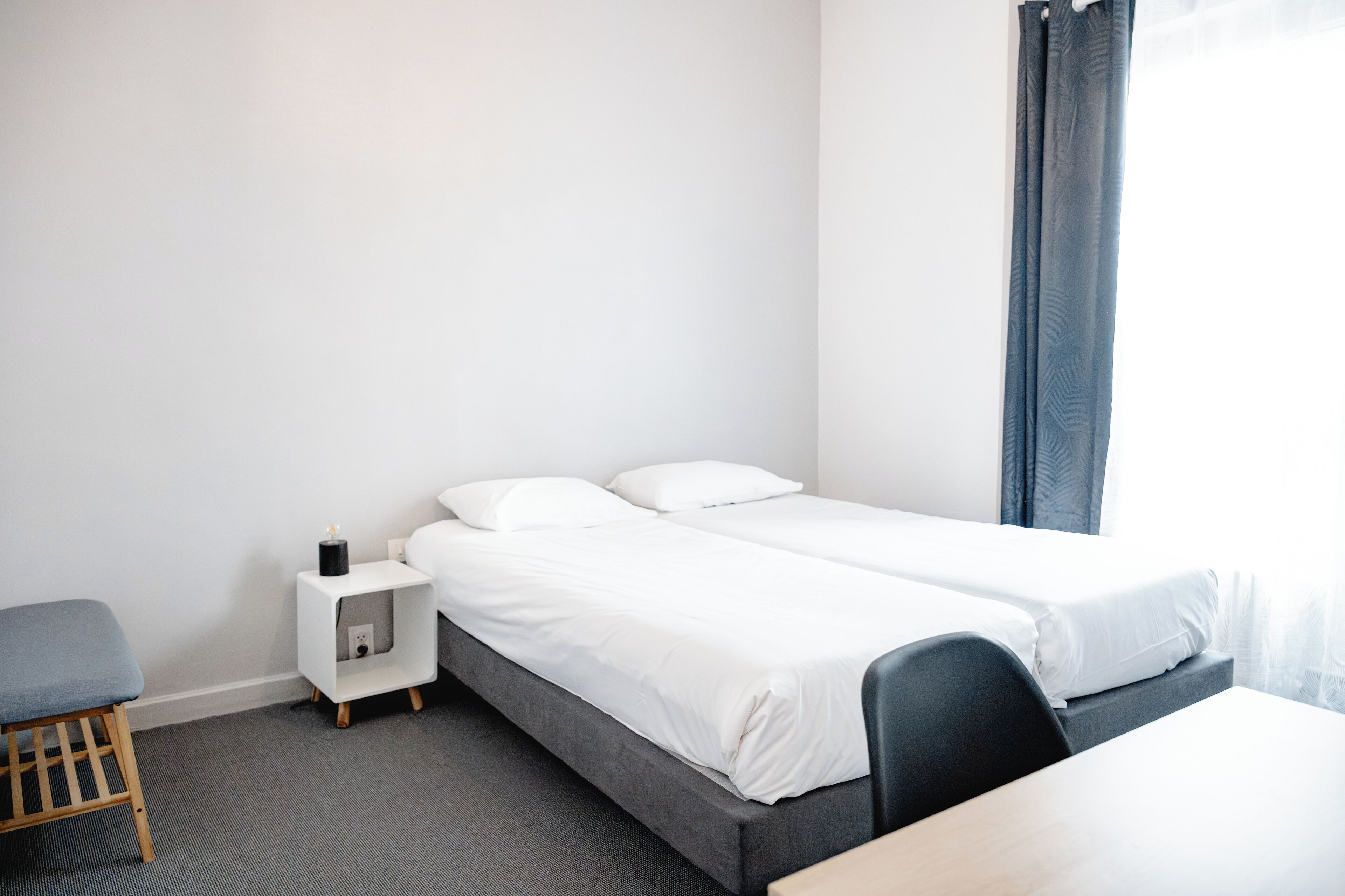 A hotel room with two single beds, a small bedside table, a chair, and a desk.