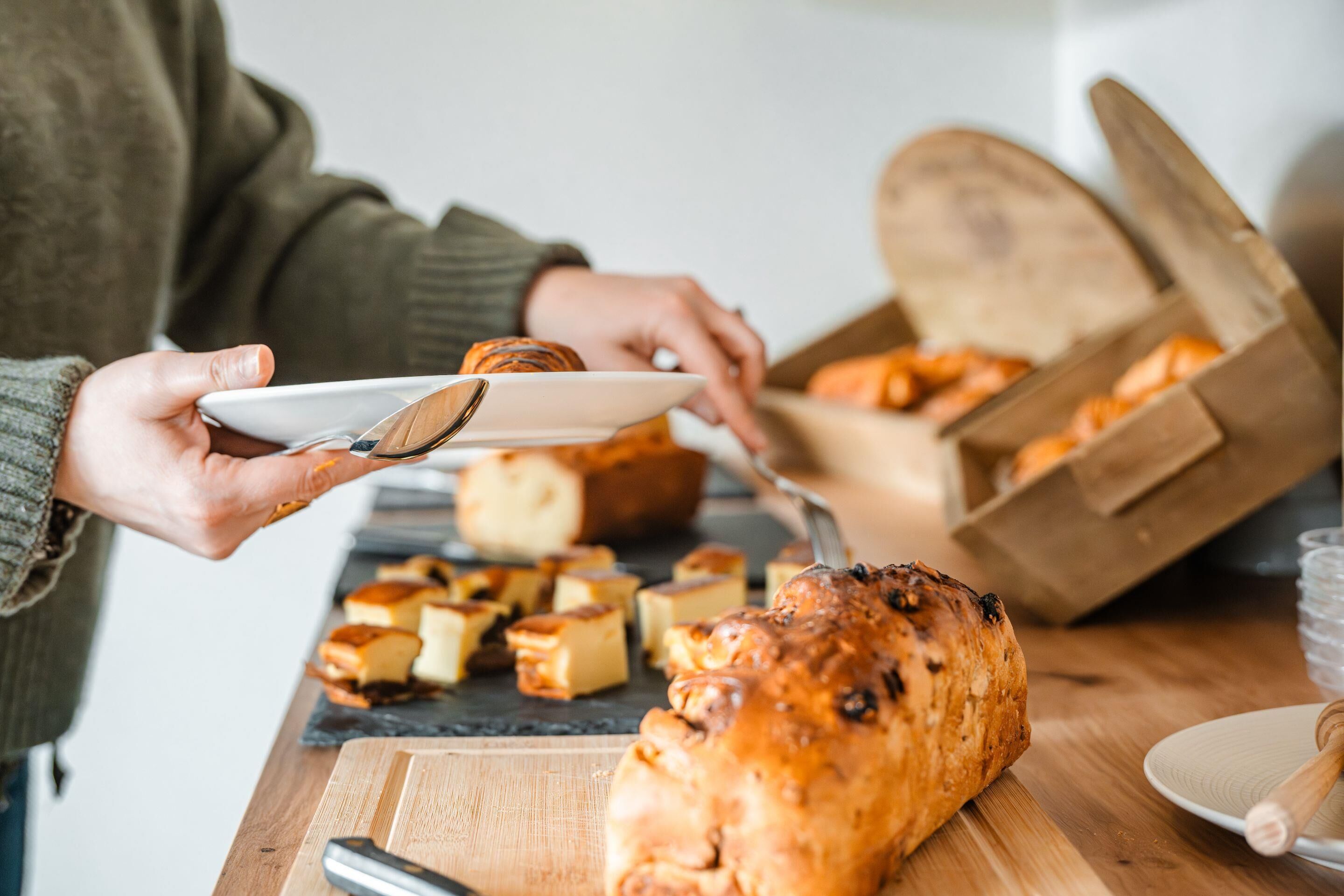 A person serving themselves food from a wooden table with a loaf of bread and various pastries.