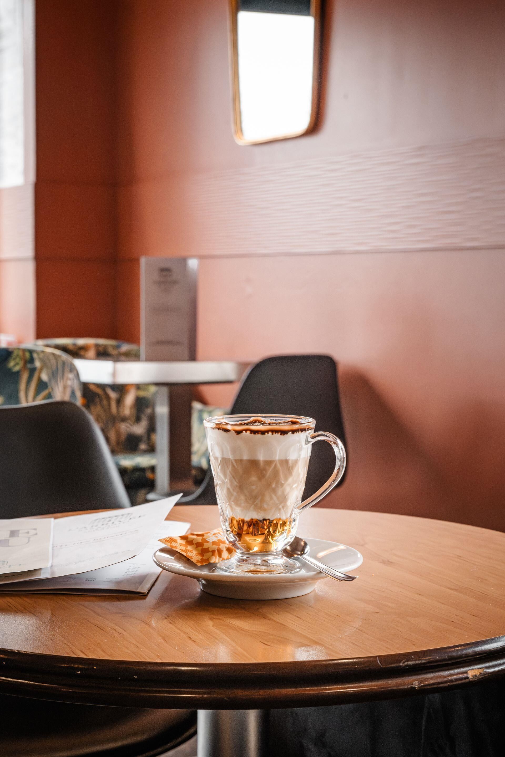 Een glazen kop koffie op een schoteltje met een lepel, op een houten tafel in een kamer met een bruine muur en een raam.
