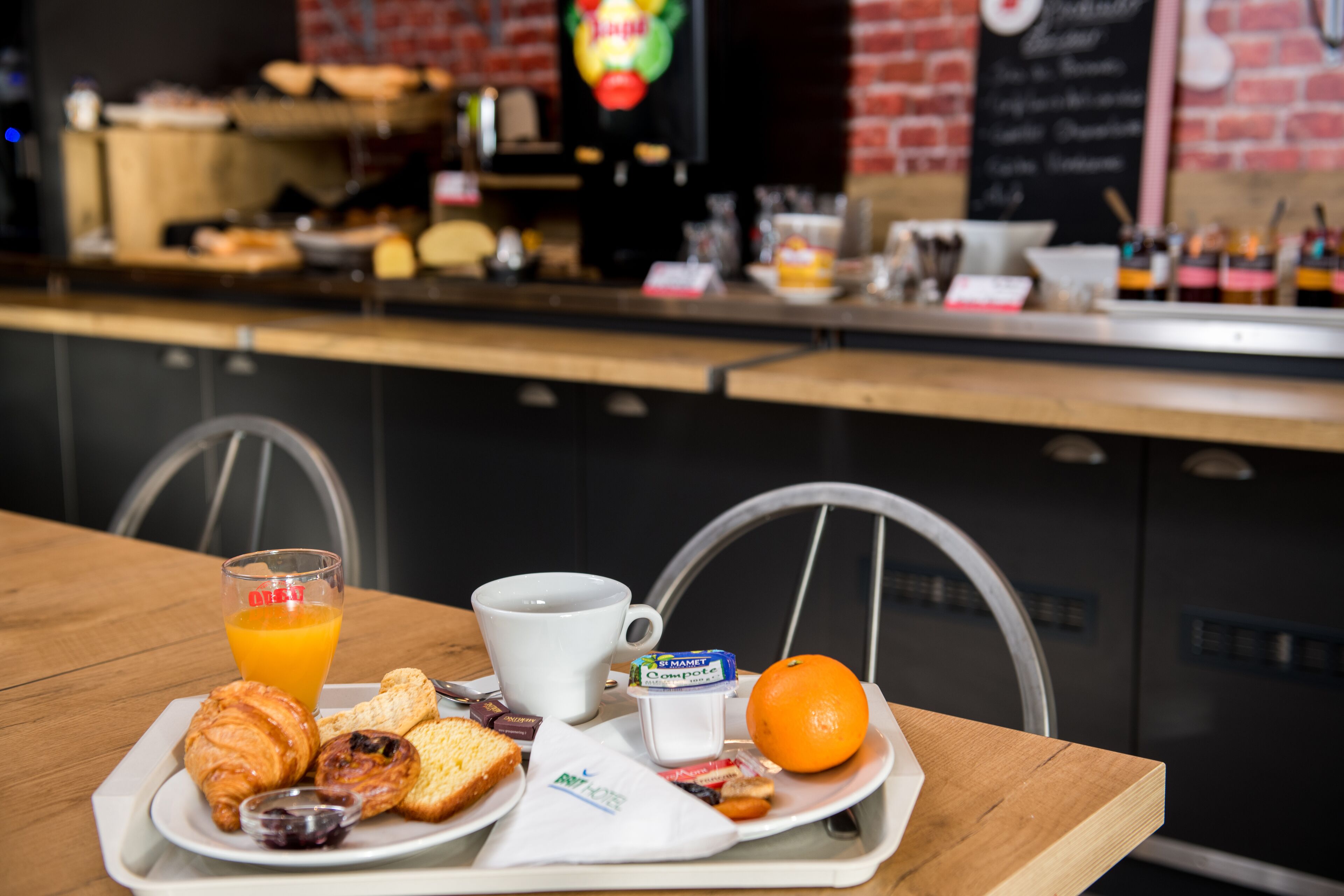 A breakfast tray with croissants, orange juice, a coffee cup, a small container of yogurt, and an orange on a wooden table in a cafe setting.