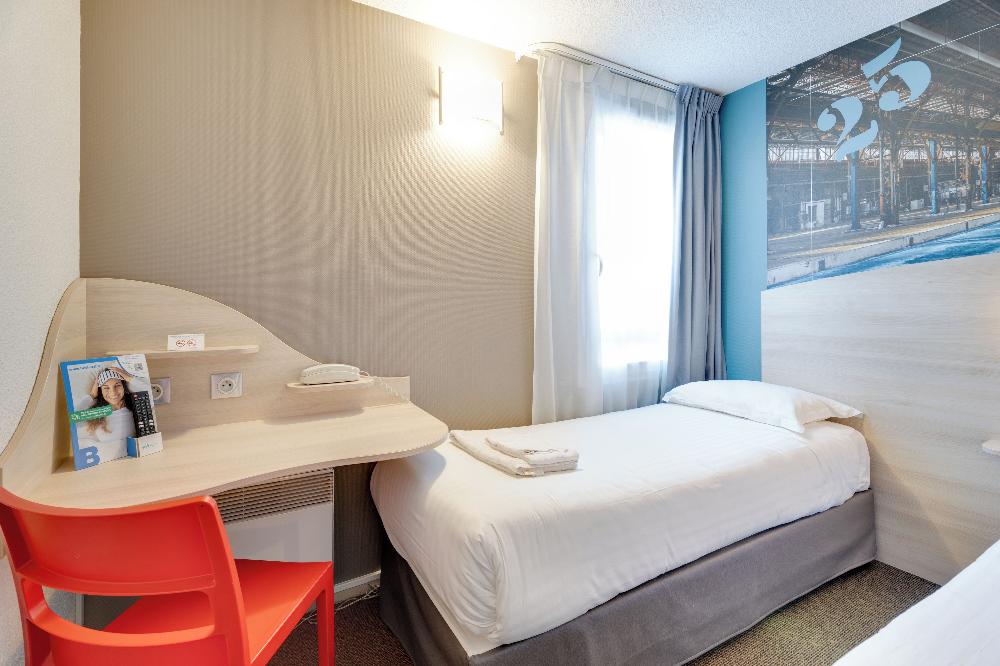 A hotel room with a bed, a desk with a chair, a telephone, and a window with curtains.