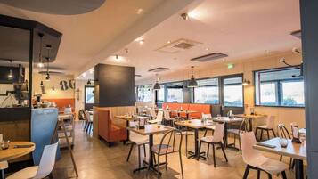 Un intérieur de restaurant moderne, doté de nombreuses tables et banquettes, de grandes fenêtres et d’un éclairage combinant lumière naturelle et artificielle.