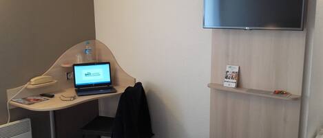 Quarto de hotel com escrivaninha, um laptop exibindo 'BRIT HOTEL', telefone, televisão e uma pequena prateleira com um folheto.