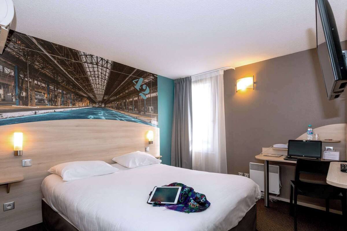 A hotel room with a bed, bedside lamps, a desk with a laptop, a water bottle, and a large mural of a bridge.