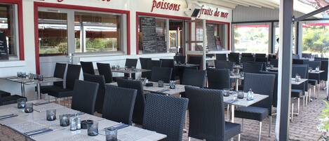 An outdoor restaurant with tables and chairs, a menu board, and a sign reading "grillades poissons fruits de mer".