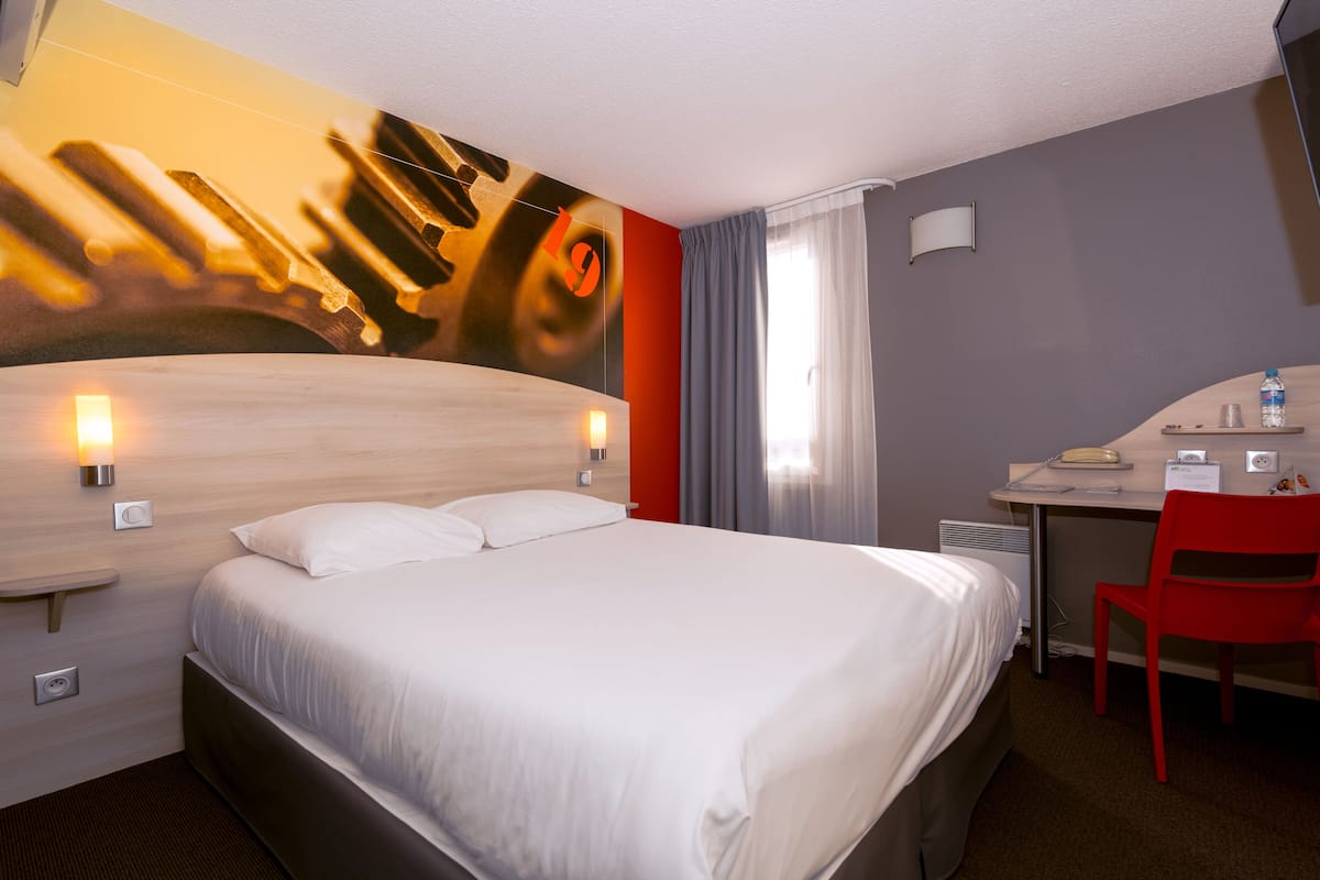 A hotel room with a large bed, a desk with a chair, and a wall-mounted lamp.