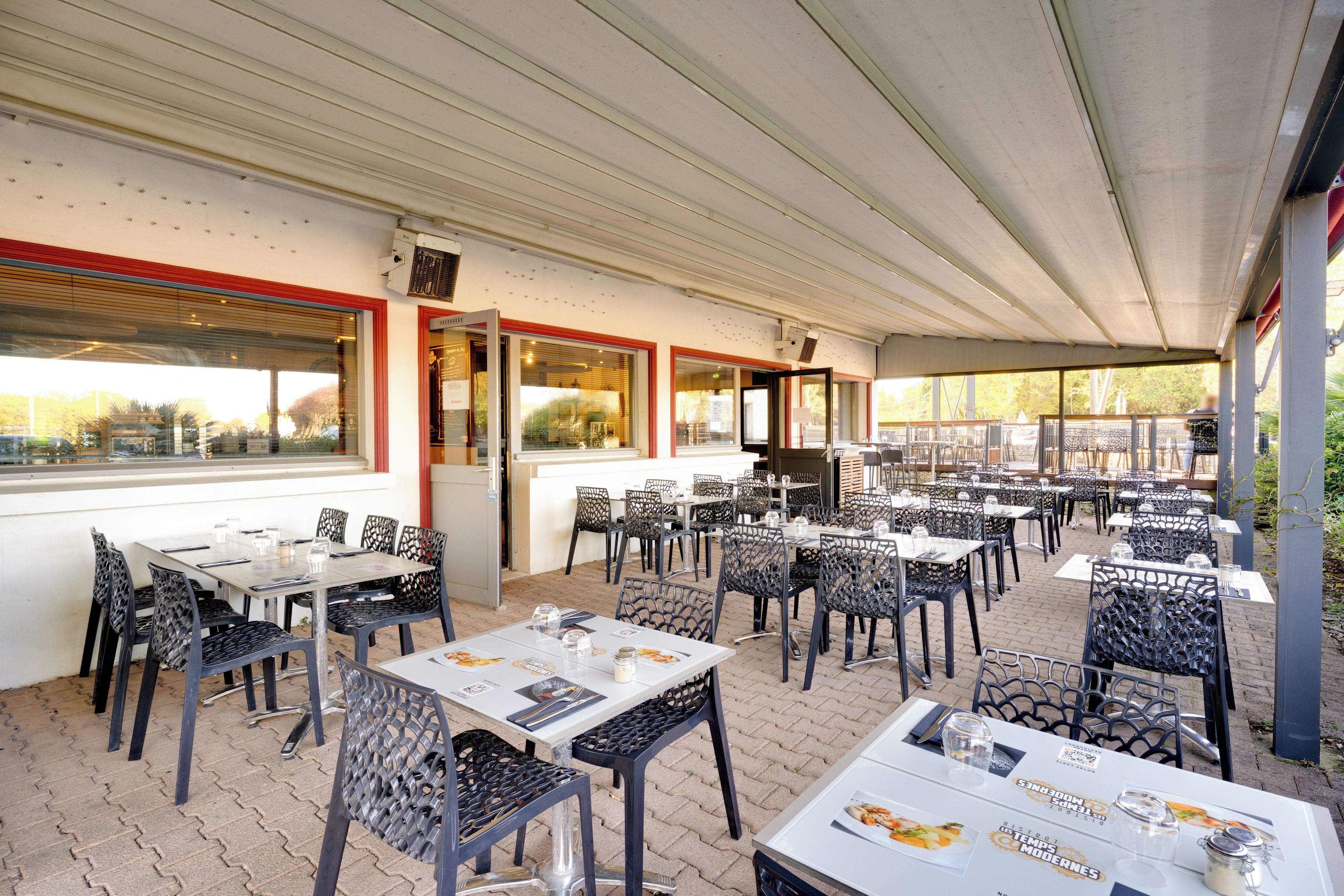 An outdoor dining area with multiple tables and chairs, covered by a roof, and large windows on one side.