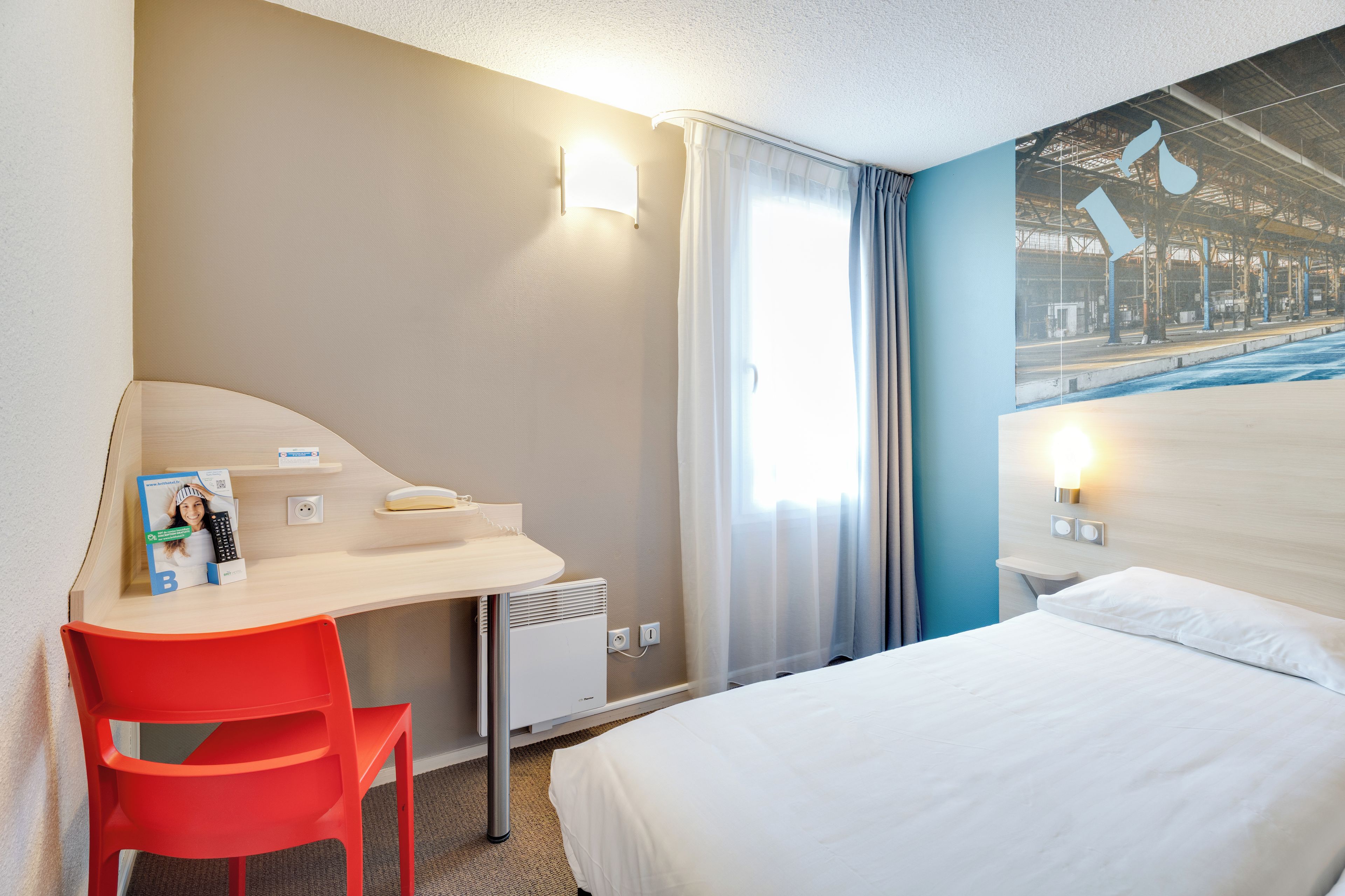Quarto de hotel com cama, escrivaninha com cadeira, janela com cortinas e parede com mural.