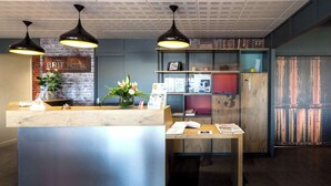 A hotel reception area with a wooden reception desk, black pendant lights, a brick wall, a flower arrangement, and a bookshelf with various items. - Brit Hotel La Rochelle Périgny (Perigny)