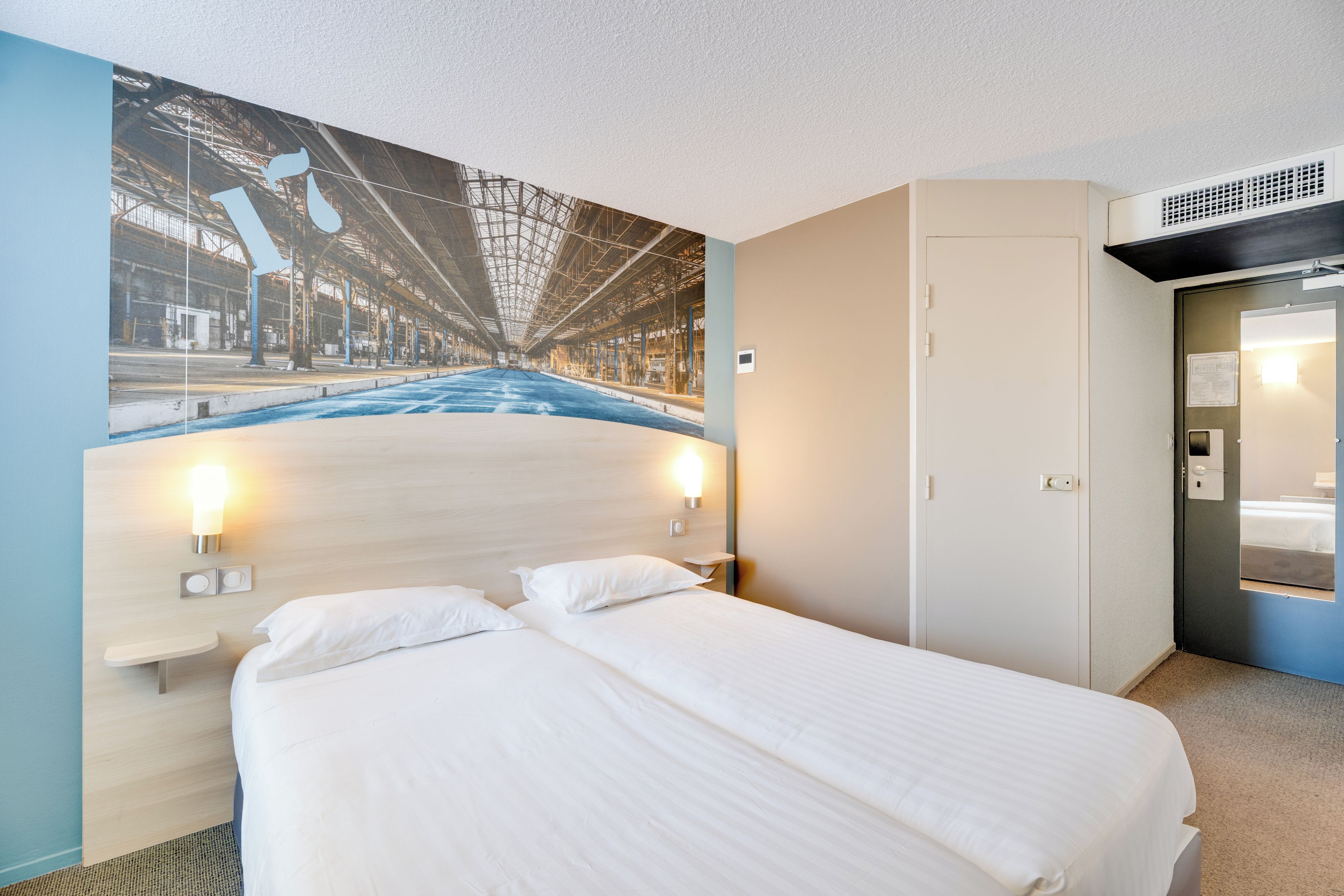 A hotel room with a bed, a wardrobe, a wall-mounted light, and a large mural of a city street.