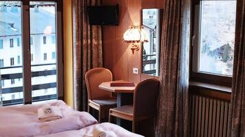 Ein Hotelzimmer mit zwei Betten, einem Schreibtisch, einem Sessel, einem Fernseher und Blick auf Gebäude durch die Fenster.
