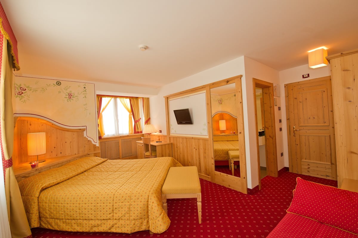 A hotel room with a large bed, a bedside table with a lamp, a small table with a chair, a mirror, and a television on the wall.