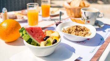 Ein Frühstückstisch mit verschiedenen Speisen, darunter Obst, eine Schüssel Müsli und eine Tasse Kaffee, draußen mit Sonnenschirmen im Hintergrund.
