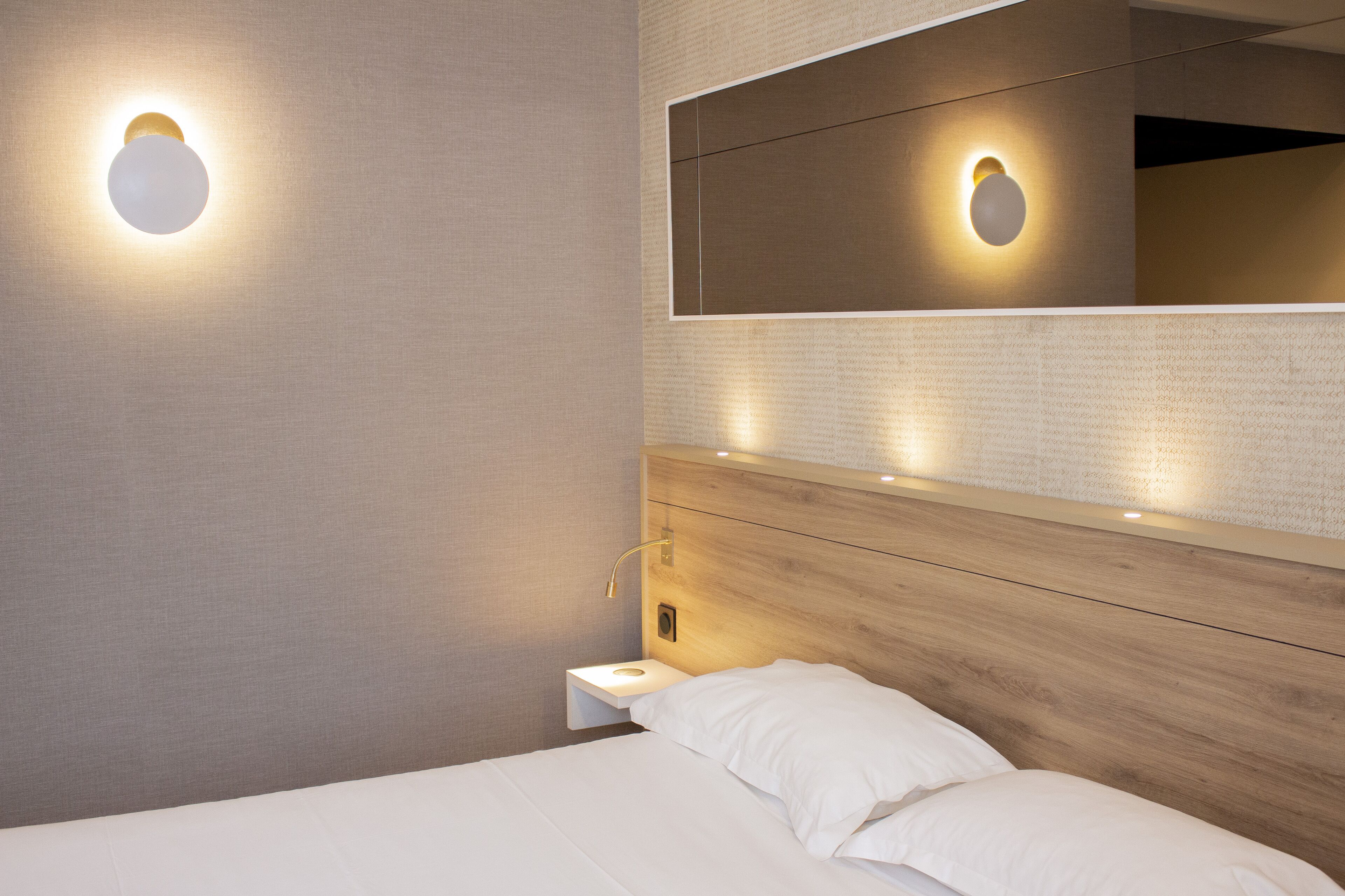 Ein modernes Hotelzimmer mit einem hölzernen Kopfteil, einem Nachttisch und einem an der Wand befestigten Spiegel.
