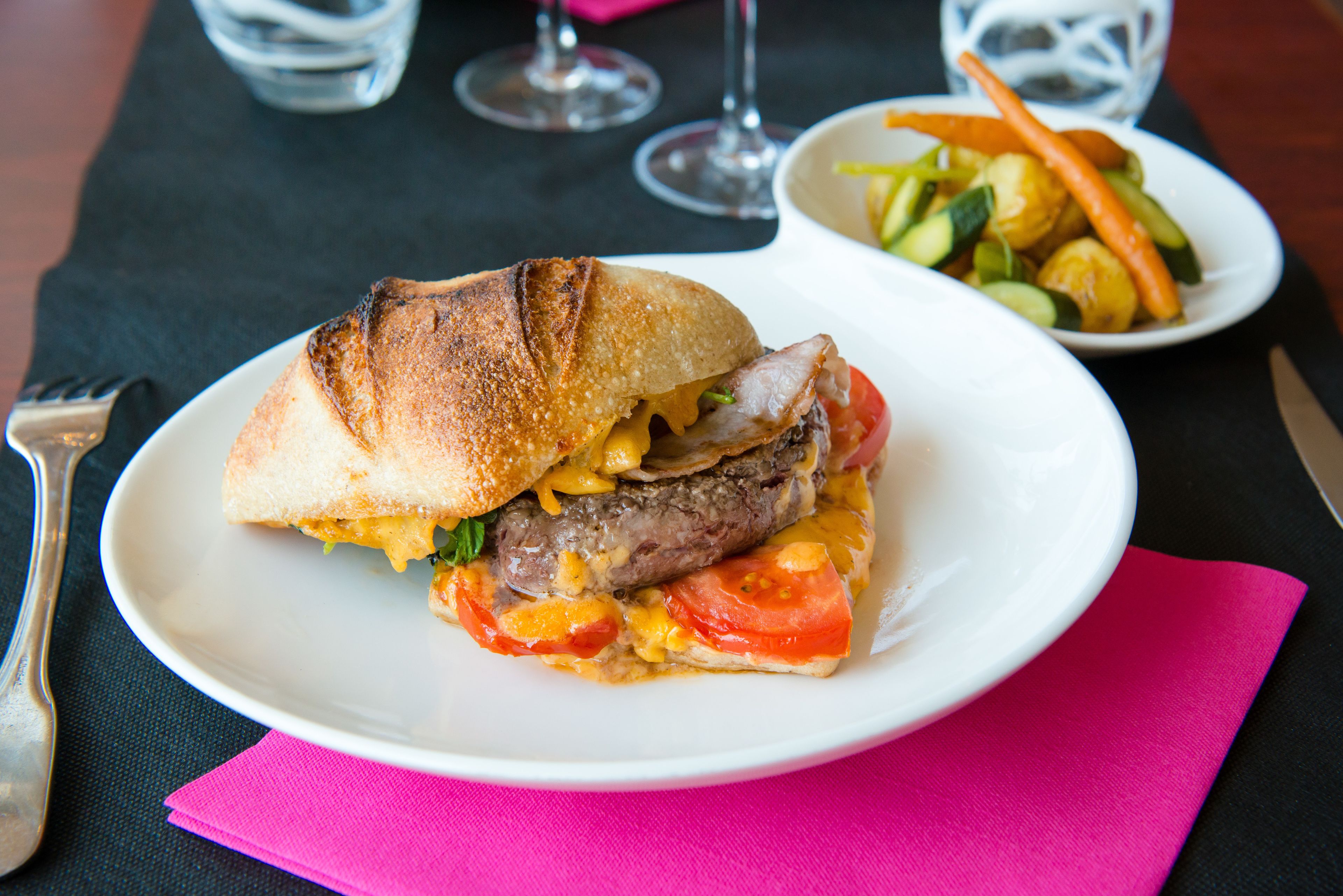 A plate with a sandwich, a bowl of vegetables, and a pink napkin on a table.