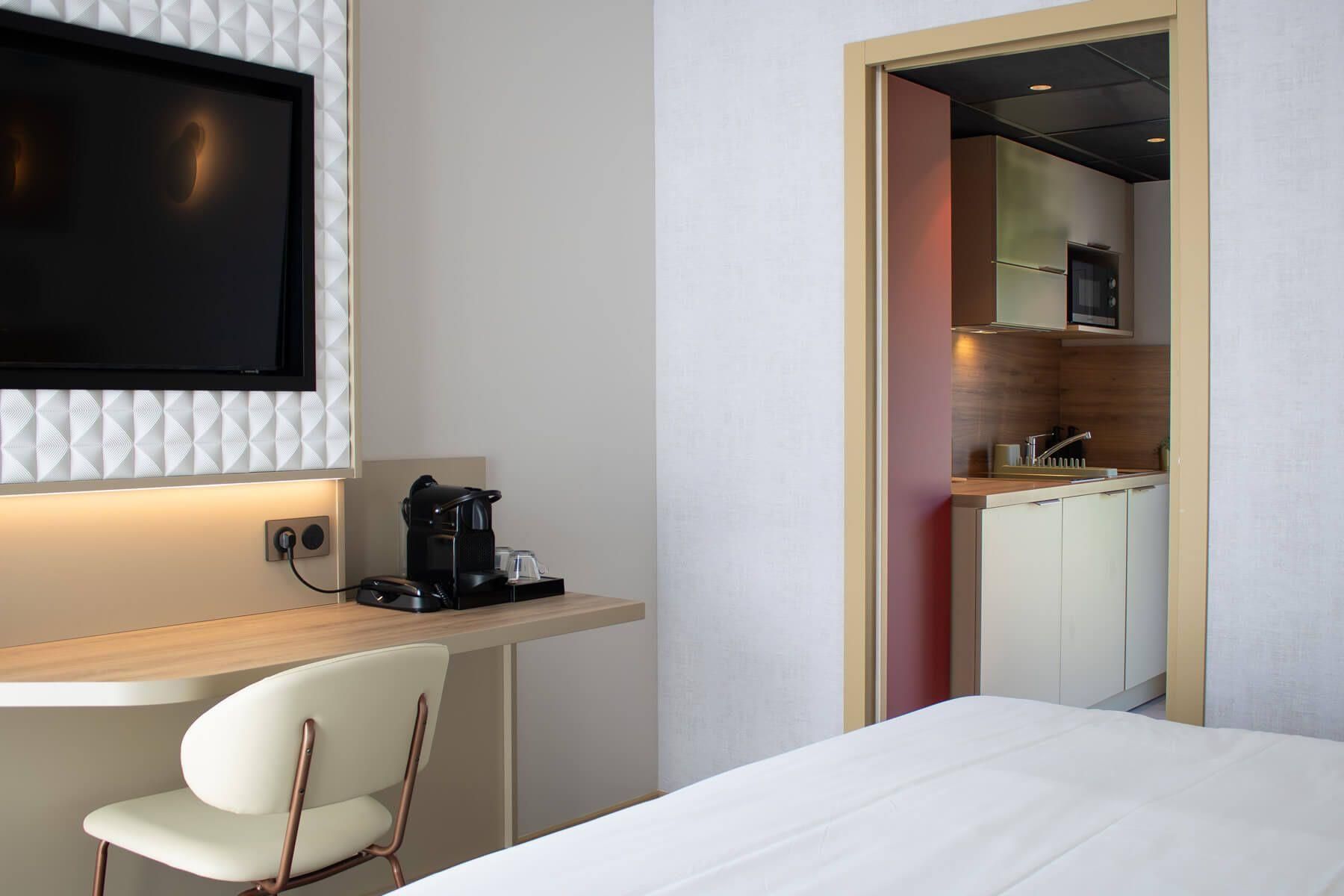 Habitación de hotel con cama, escritorio con cafetera, televisor montado en la pared y vista al área de la cocina.