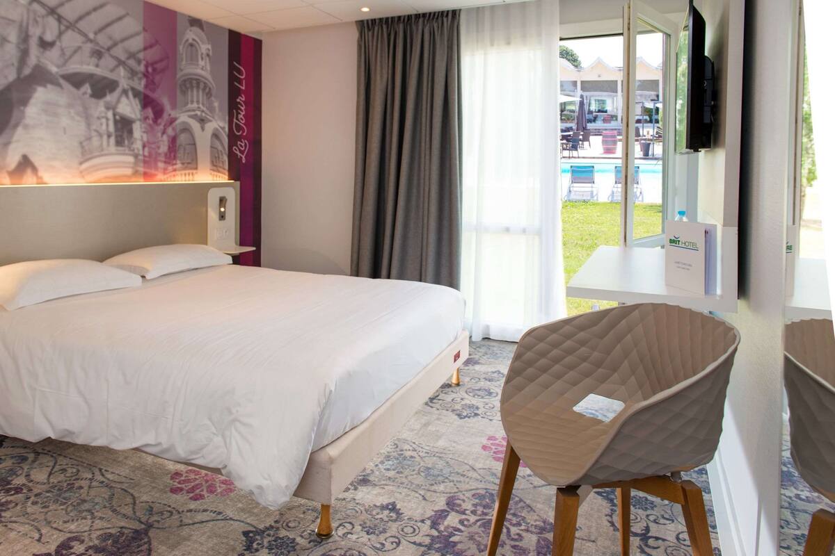 Ein Hotelzimmer mit einem großen Bett, einem Sessel, einem Fernseher und Blick ins Freie.