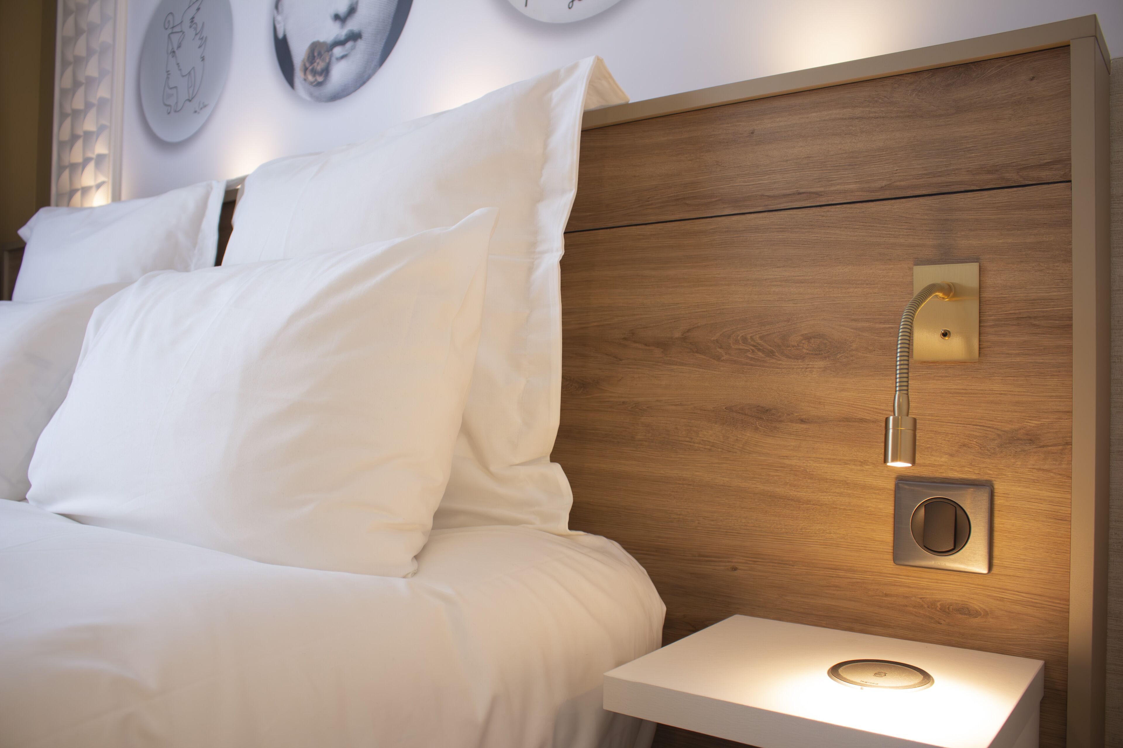 Un lit bien fait, avec du linge de lit blanc, une tête de lit en bois, une table de chevet avec une lampe et un luminaire fixé au mur.