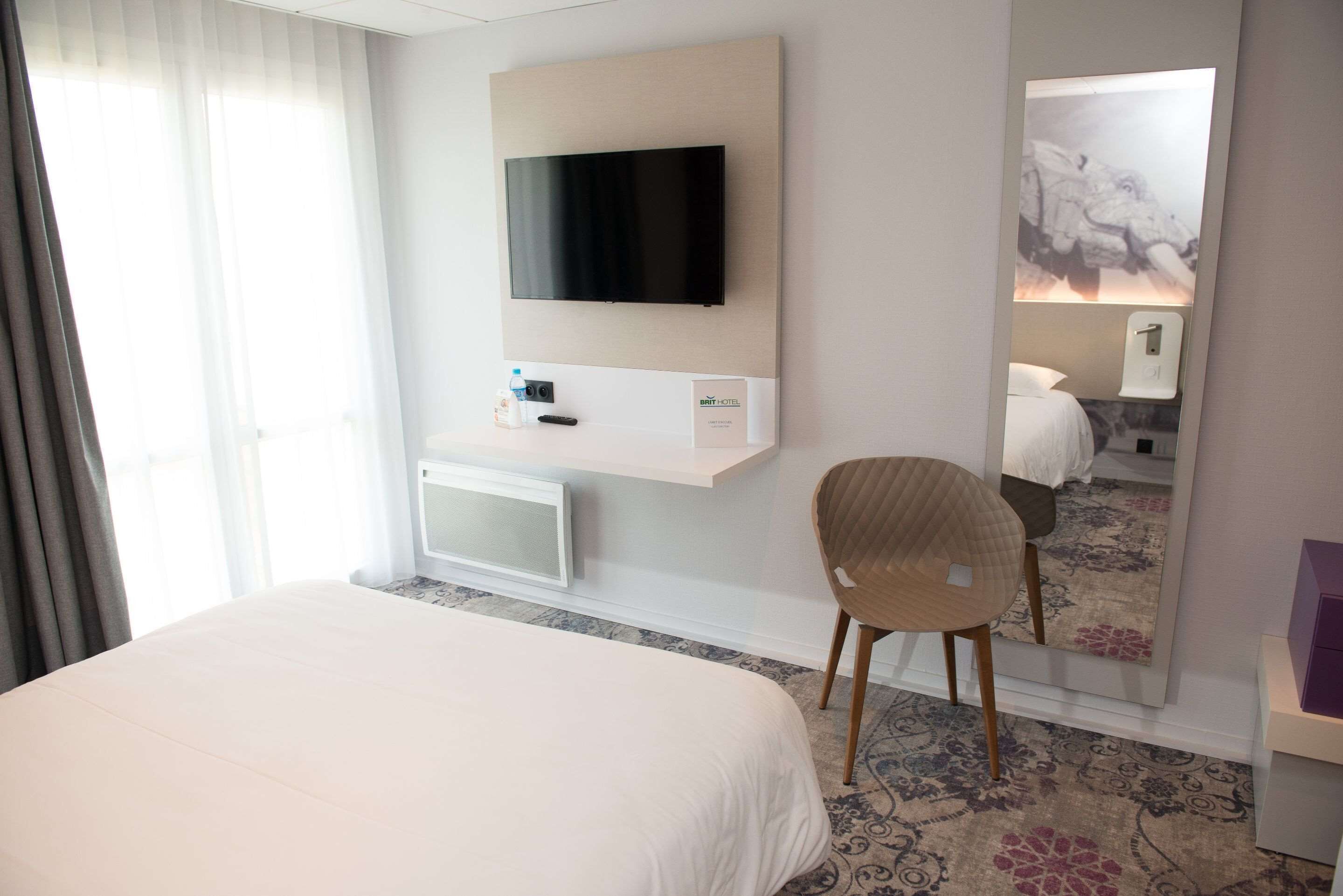 A hotel room with a large bed, a flat-screen TV mounted on the wall, a small table with a bottle and remote, a chair, and a mirror reflecting the room.