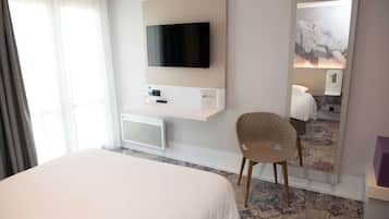 A hotel room with a large bed, a flat-screen TV mounted on the wall, a small table with a bottle and remote, a chair, and a mirror reflecting the room.