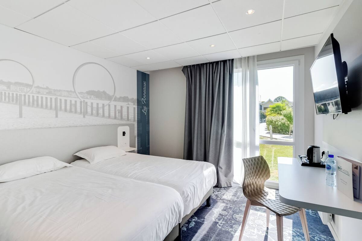 Ein Hotelzimmer mit zwei Betten, einem Schreibtisch, einem Sessel, einem Fernseher und einem Fenster mit Ausblick.
