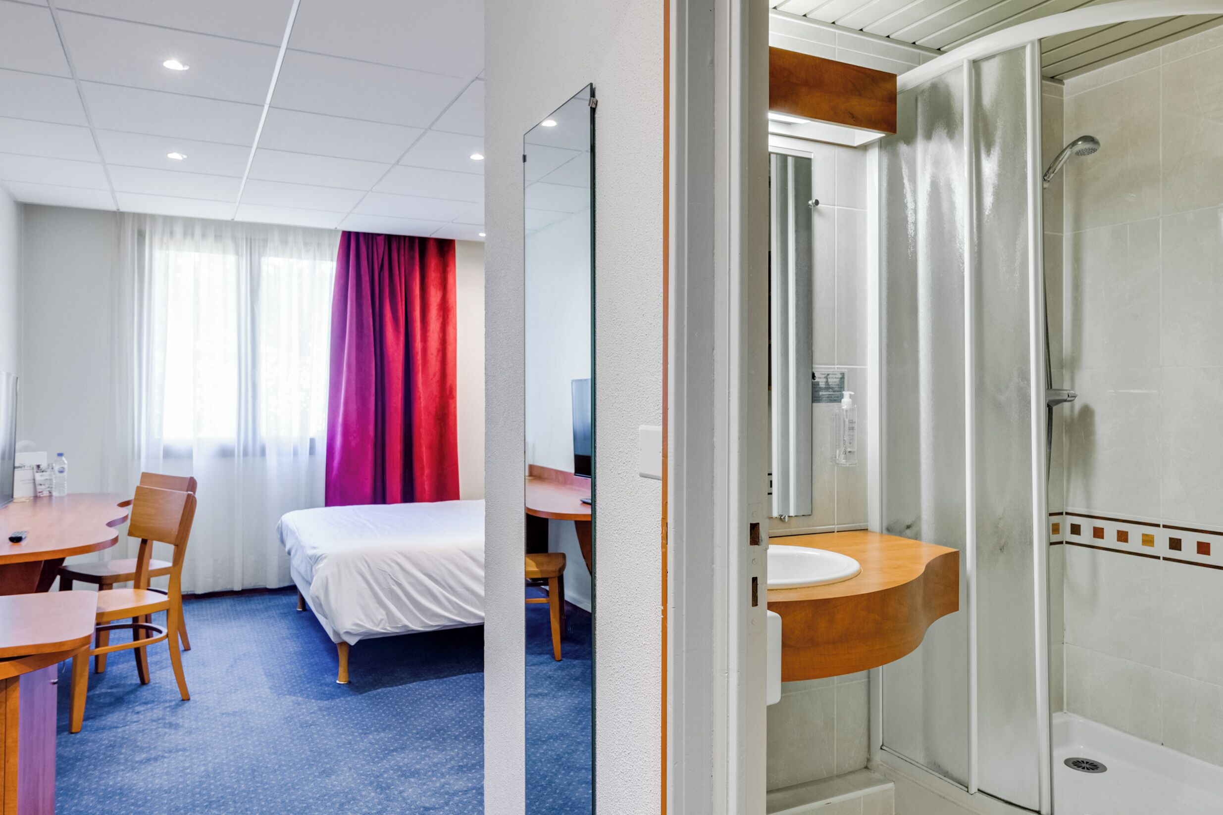 Habitación de hotel con cama, escritorio, silla, baño con ducha y espejo.