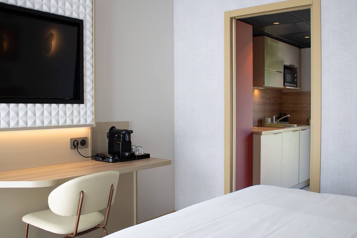 Ein Hotelzimmer mit Bett, Schreibtisch mit Kaffeemaschine, Wand-Fernseher und Blick in den Küchenbereich.