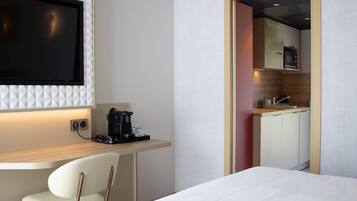 A hotel room with a bed, a desk with a coffee maker, a TV mounted on the wall, and a view into the kitchen area.