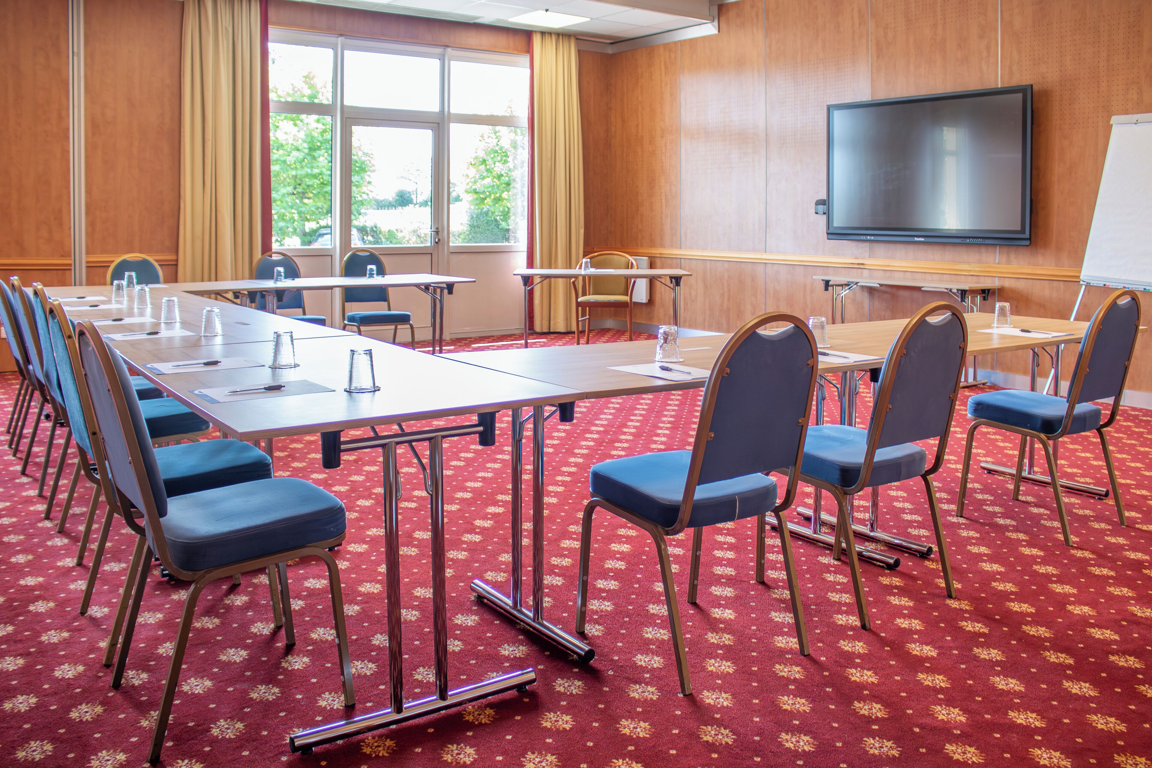Sala de conferência com uma mesa comprida, cadeiras, tela de projetor e piso coberto por um tapete vermelho.