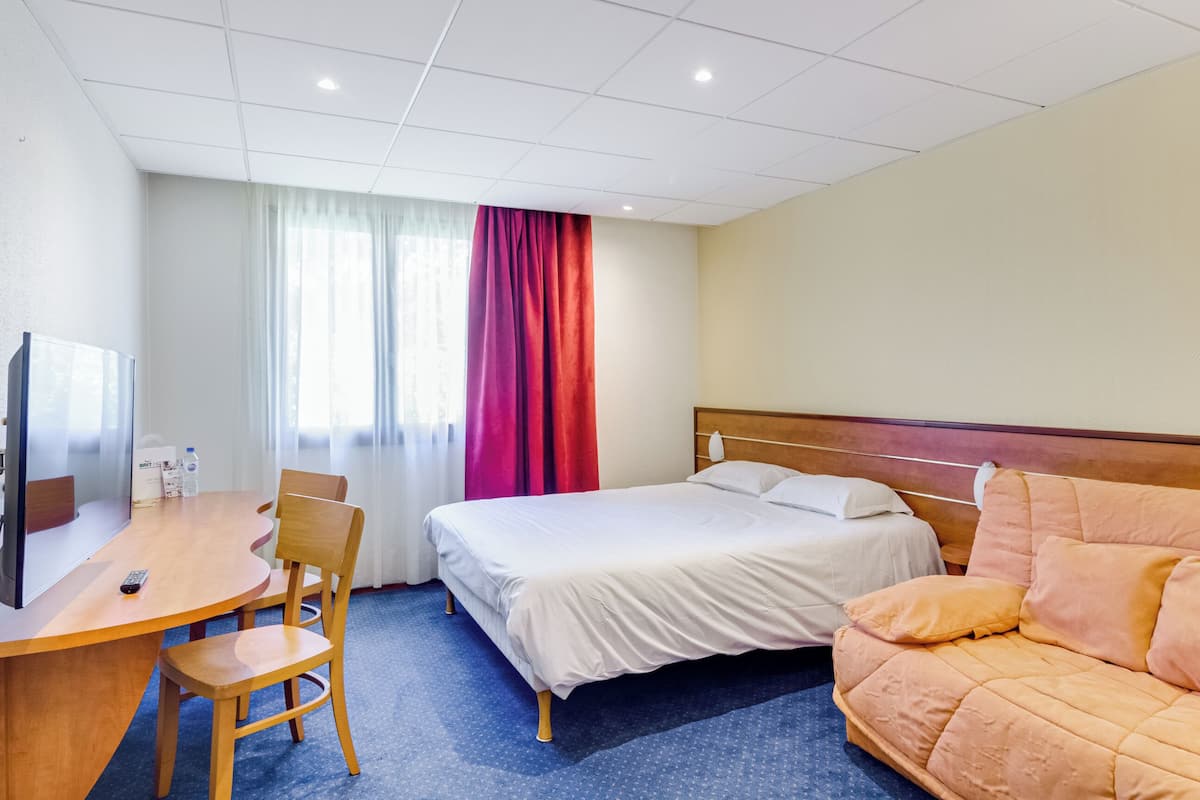 Ein Hotelzimmer mit Bett, einer Couch, einem Schreibtisch mit Stuhl, einem Fernseher und einem Fenster mit Vorhängen.