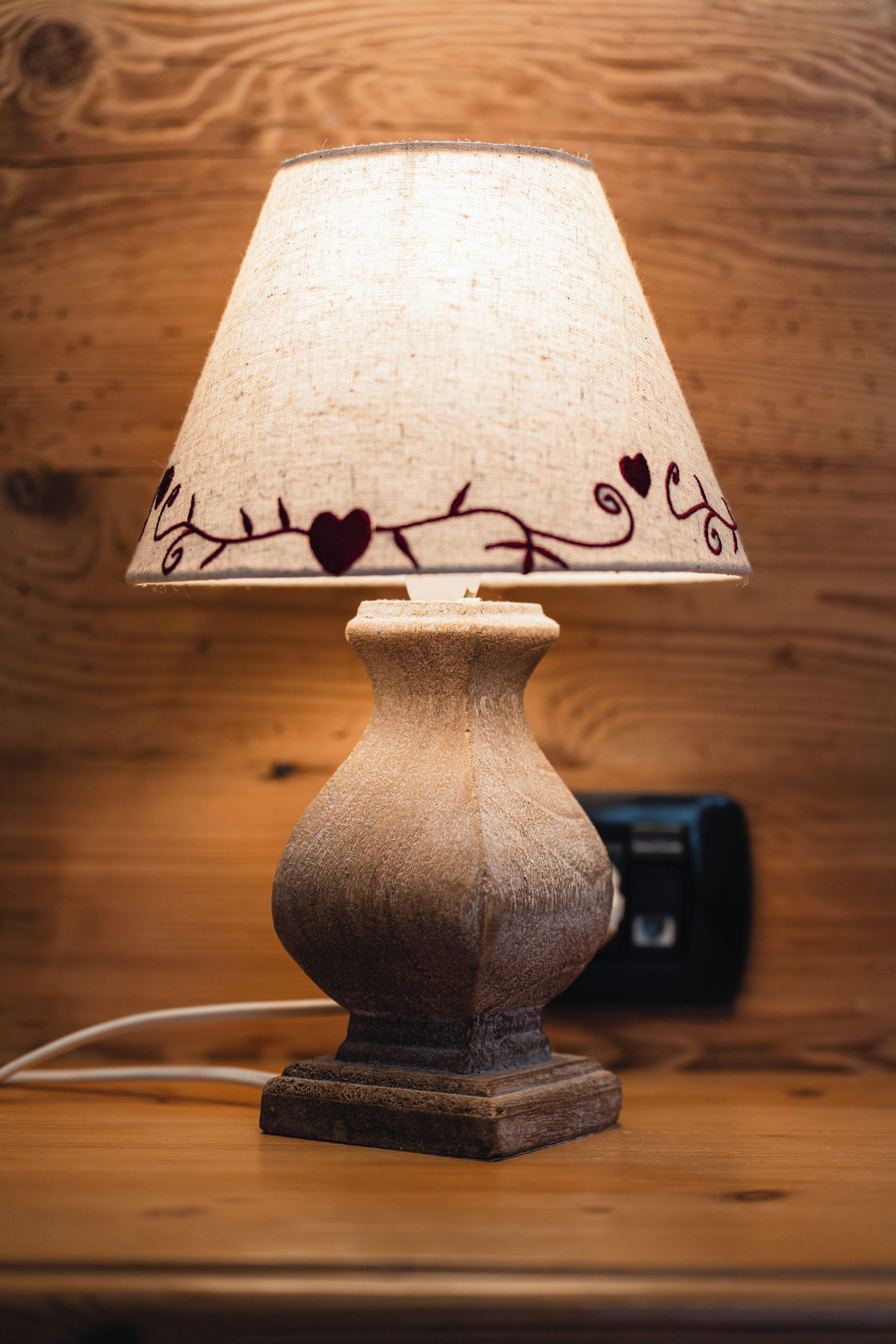 Una lampada da tavolo con un paralume strutturato, decorato con motivi a cuore e tralci, posata su una superficie in legno.