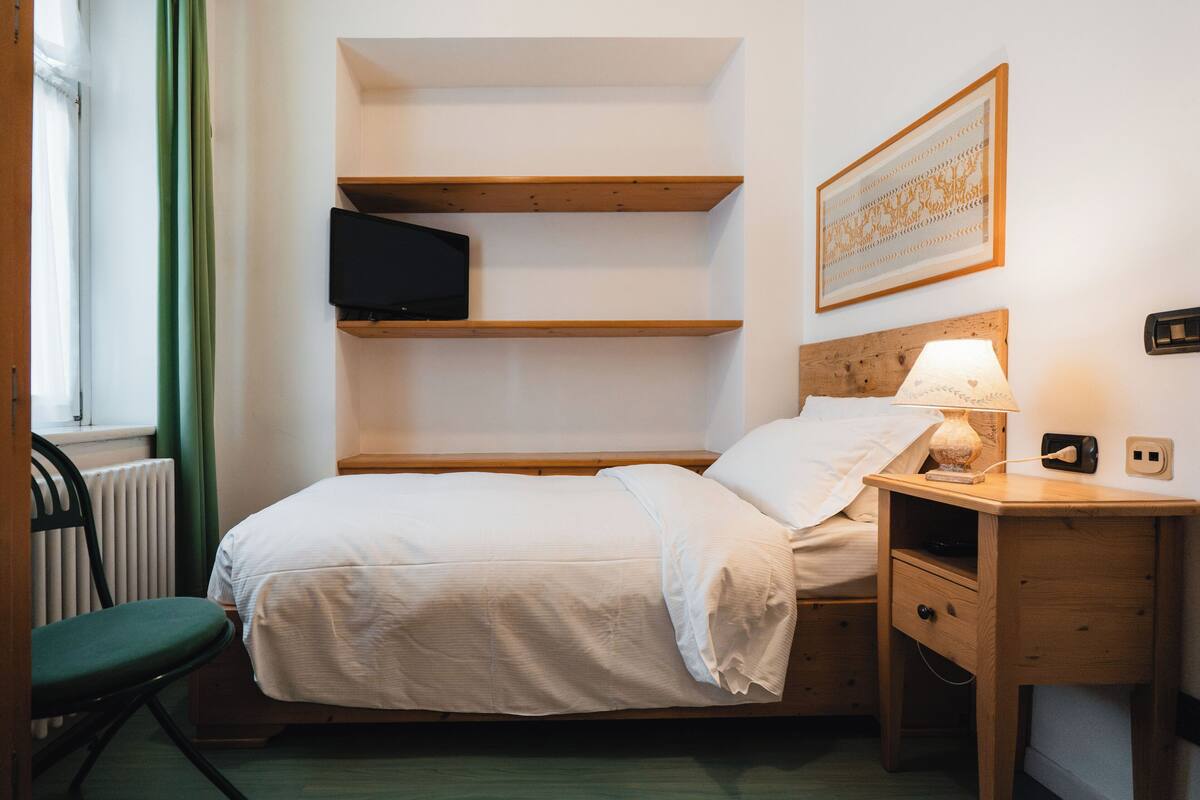 A neatly arranged bedroom with a bed, a bedside table with a lamp, a TV on a shelf, and a framed picture on the wall.