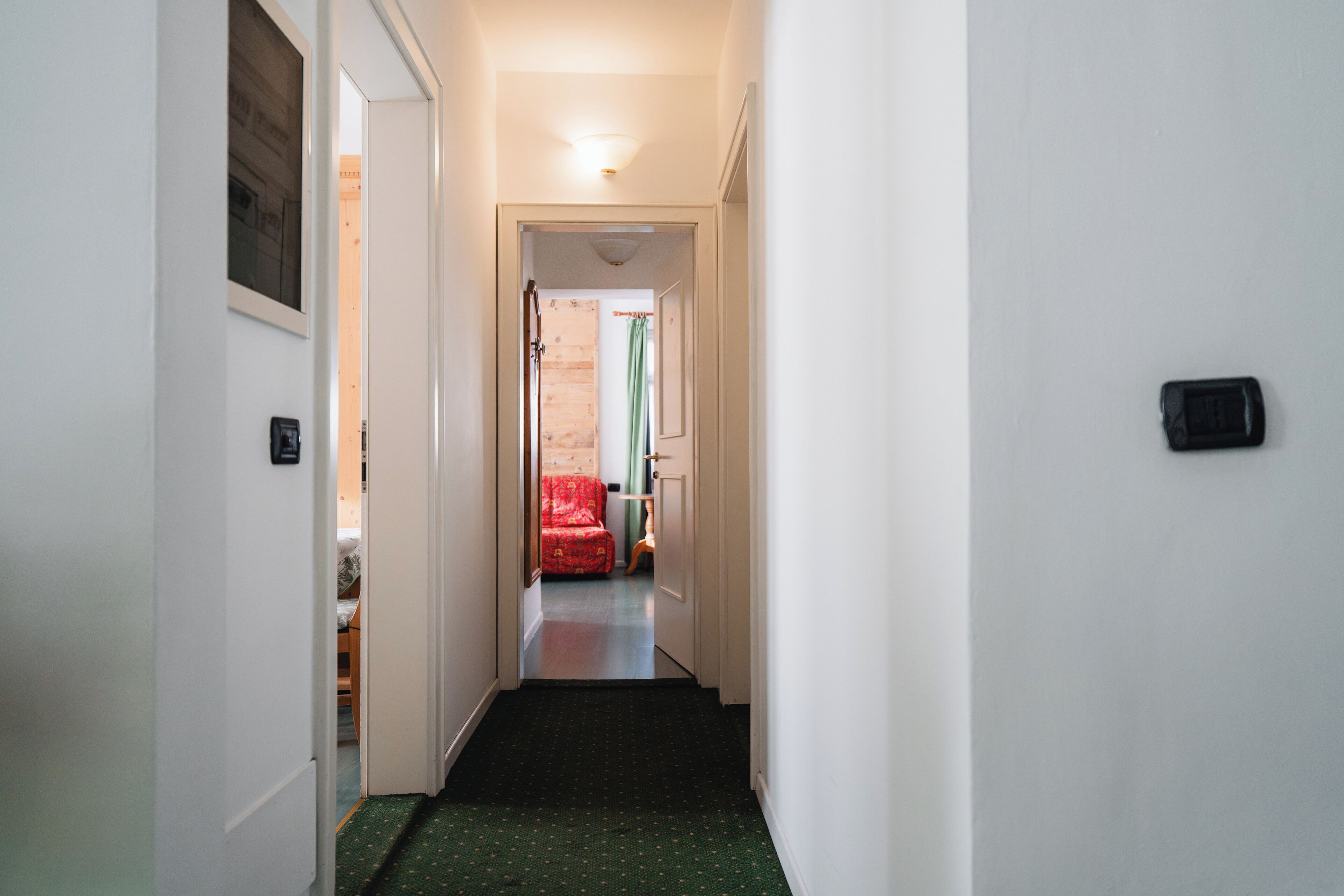A narrow hallway with a green carpet leading to a room with a red chair and a bed.