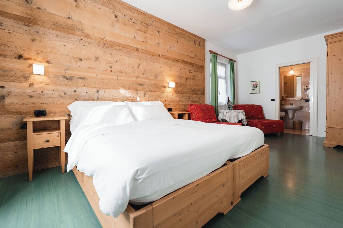 A wooden bed in a room with a green floor, a nightstand, a red chair, and a wooden wardrobe.