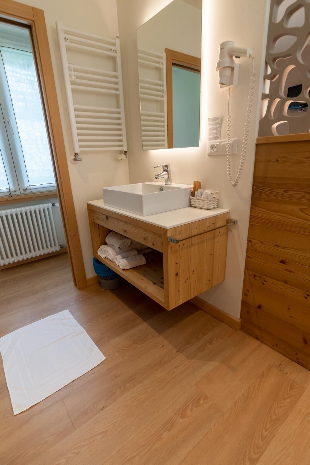 Un baño con tocador de madera, lavamanos blanco y un espejo. Hay un perchero para toallas y una alfombra de baño blanca en el suelo.