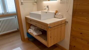 Un baño con tocador de madera, lavamanos blanco y un espejo. Hay un perchero para toallas y una alfombra de baño blanca en el suelo.