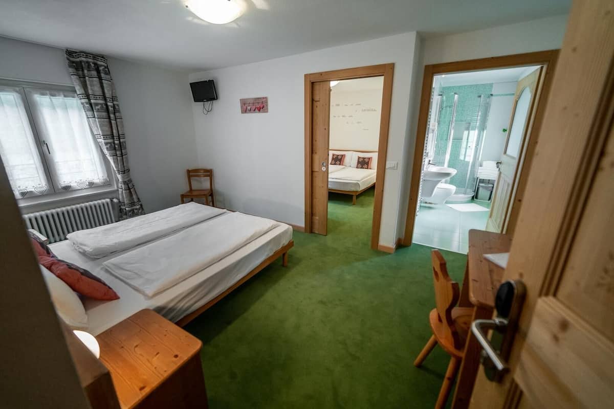 Un dormitorio con cama doble, una silla de madera, un escritorio, un radiador, televisión y un baño visible a través de una puerta abierta.